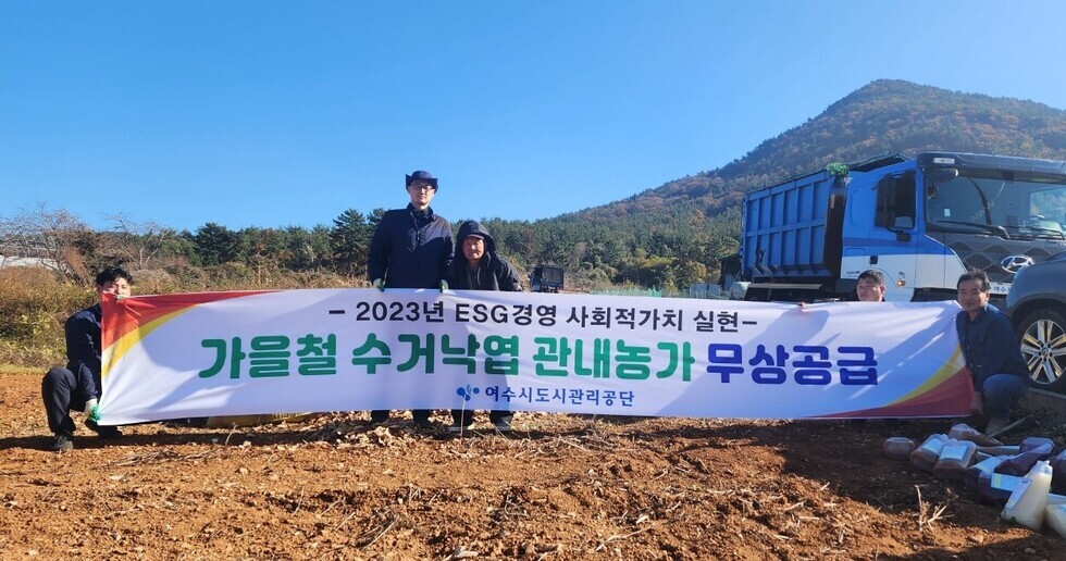 흙 땅 위에 2023년 ESC 경영 사회적 가치 실현 / 가을철 수거낙엽 관내농가 무상공급 / 여수시도시관리공단 현수막 뒤에 사람들이 서 있는 모습