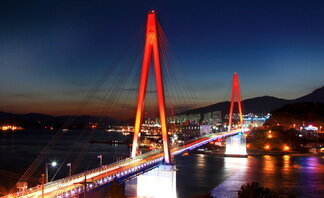 붉은 조명이 아름답게 비춰진 돌산대교의 야경. 다리는 푸른 바다 위를 가로지르며 도시의 불빛과 어우러져 화려한 분위기를 자아낸다. 배경에는 어두운 하늘과 산자락이 어렴풋이 보이며, 다리 아래 수면에는 조명빛이 반사되어 반짝인다.