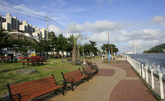 종포해양공원 산책로의 모습. 오른쪽으로는 푸른 바다가 펼쳐져 있고, 해안선을 따라 하얀 난간과 붉은 벽돌길이 이어진다. 왼쪽에는 나무 벤치와 잔디밭이 있고, 그 너머로 아파트 단지가 보인다. 멀리에는 다리가 보이며, 전체적으로 맑은 하늘과 구름이 어우러져 여유롭고 평화로운 분위기를 자아낸다.