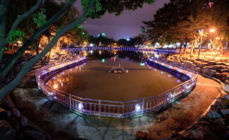 거북선 공원의 야경. 중앙에 조용한 연못이 자리하고 있으며, 연못 주변은 울타리와 보라색 조명으로 아름답게 장식되어 있다. 양옆으로는 키 큰 나무들이 우거져 있고, 오른쪽 산책로에는 가로등 불빛이 따뜻하게 공원을 비추고 있다. 조용하고 낭만적인 분위기 속에서 도시의 불빛이 멀리 반짝인다.