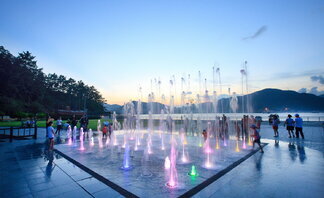 오동도의 여름 저녁, 분수광장에서 알록달록한 조명이 켜진 분수들이 시원하게 솟구치고 있다. 분수 주위에는 아이들과 가족들이 모여 시원한 물놀이를 즐기고 있고, 일부 사람들은 주변 벤치와 바다를 배경으로 풍경을 감상하고 있다. 배경에는 석양이 지는 하늘과 산, 바다가 어우러져 고요하고 아름다운 분위기를 자아낸다.