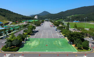 진남체육공원의 전경이 한눈에 들어오는 사진. 전면에는 초록색 농구장이 위치해 있고, 그 뒤로는 넓은 주차장이 이어진다. 양쪽에는 푸르른 나무들과 산이 펼쳐져 있으며, 오른쪽에는 축구장이 보인다. 멀리에는 도심과 아파트, 푸른 산들이 조화를 이루고 있다. 하늘은 맑고 파랗게 개어 있어 쾌청한 날씨를 보여준다.