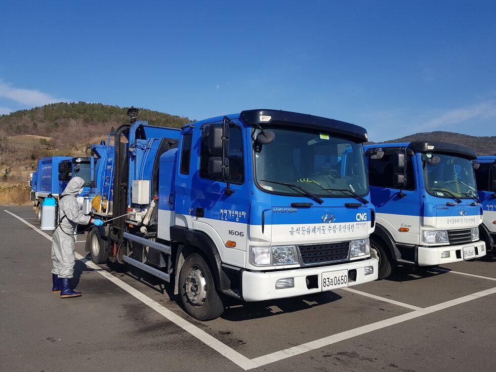 파란색 음식물류 폐기물 수거 차량이 주차된 공간에서 회색 방역복을 착용한 남성 작업자가 차량 옆면과 장비를 소독하고 있다. 차량 앞면에는 '음식물류폐기물 수집·운반전용차량'이라는 문구가 적혀 있으며, 같은 디자인의 차량들이 줄지어 서 있다.