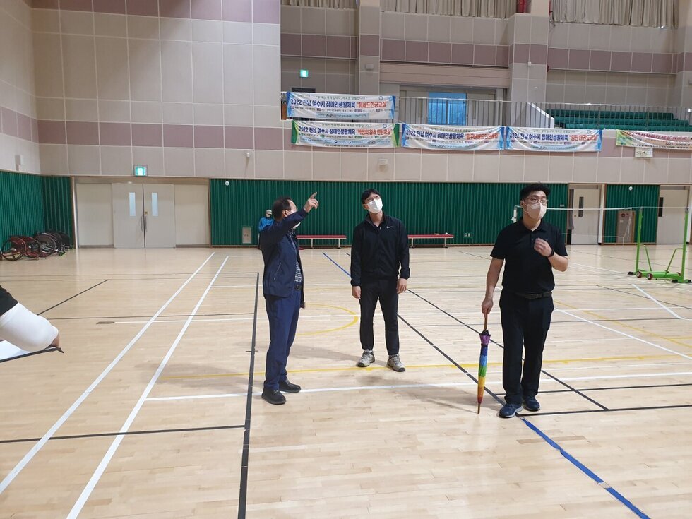 실내 체육관에서 마스크를 착용한 세 명의 남성이 천장을 바라보며 시설을 점검하고 있다. 한 명이 손가락으로 위쪽을 가리키며 설명하고 있고, 나머지 두 명은 고개를 들어 해당 방향을 주의 깊게 살피고 있다. 바닥에는 배드민턴 라인이 그려져 있고, 한 남성은 무지개색 우산을 들고 있다. 배경에는 행사 현수막이 걸려 있으며 전체적으로 점검 중인 진지한 분위기이다.