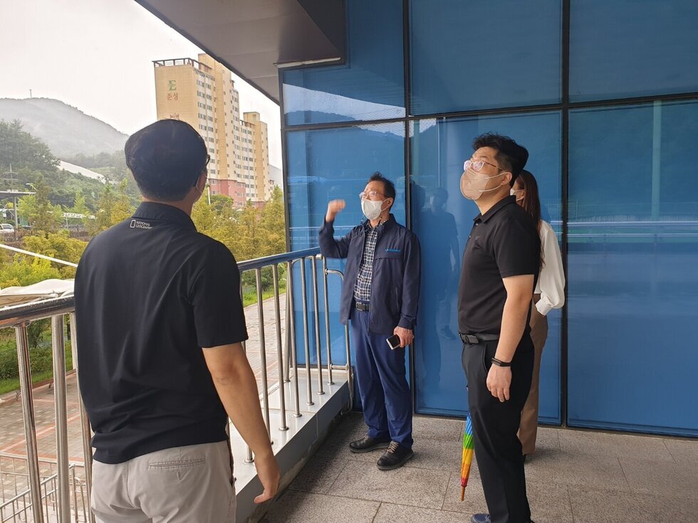 건물 외부 베란다에서 네 명의 사람들이 마스크를 착용한 채 시설을 점검하고 있다. 한 남성이 천장을 가리키며 설명하고 있고, 나머지 사람들은 같은 방향을 바라보며 집중하는 모습이다. 뒤쪽에는 파란색 유리벽과 안전 난간이 있으며, 배경으로는 산과 아파트 단지가 보인다. 우산을 든 사람도 있어 비가 오거나 흐린 날씨로 보인다.