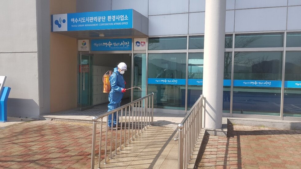 여수시도시관리공단 환경사업소 건물 출입구 앞에서 파란색 방역복과 흰색 헬멧을 착용한 남성 작업자가 분무기를 이용해 소독 작업을 하고 있다. 등에 소독약 통을 메고 있으며, 출입로 난간과 바닥을 중심으로 방역 중이다.