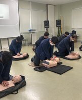 8명의 사람들이 강의실 바닥에 무릎을 꿇고 CPR 마네킹을 대상으로 심폐소생술 교육을 받고 있다. 각자 앞에 놓인 마네킹에 압박을 가하며 실습 중이며, 뒤편 스크린에는 강사가 CPR 시범을 보이는 영상이 재생되고 있다. 참가자들은 동일한 네이비 작업복을 착용하고 있으며 마스크를 착용하고 있다.