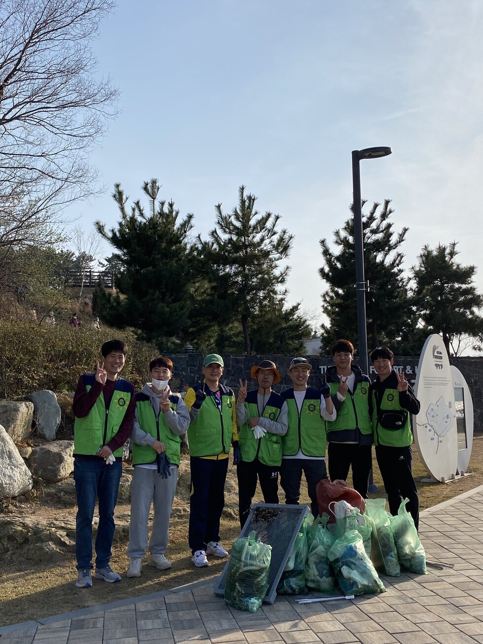 초록색 조끼를 입은 20대 ~ 40대 남성 자원봉사자 7명이 쓰레기봉투와 분리수거물 옆에 나란히 서서 밝게 웃고 있다. 모두 손으로 브이(V) 포즈를 취하고 있으며, 봉사 활동 후 정리된 여러 개의 쓰레기봉투와 그물망이 바닥에 놓여 있다. 배경에는 나무와 바위, 약간의 오르막길이 보이며, 맑은 날씨 속 따뜻한 분위기가 느껴진다. 
