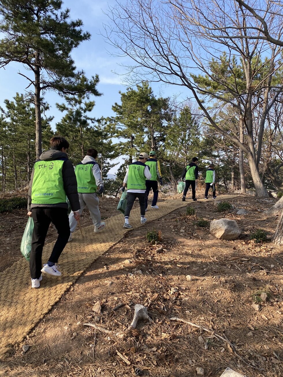 연두색 조끼를 입은 자원봉사자 6명이 숲속 산책로를 따라 걸으며 환경정화 활동을 하고 있다. 모두 쓰레기봉투와 집게를 손에 들고 있다. 나무가 울창한 숲길 사이로 햇빛이 비추고, 참가자들은 뒷모습을 보이고 있으며 일렬로 길을 따라 이동 중이다.