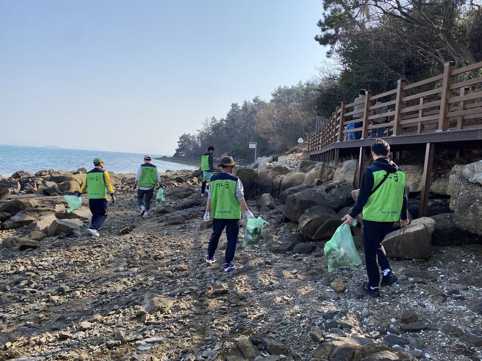 연두색 자원봉사 조끼를 입은 5명의 참가자들이 해변의 바위길을 따라 걸으며 해안 정화 활동을 하고 있다. 모두 손에 초록색 쓰레기봉투를 들고 있으며  오른쪽에는 나무 데크가 설치되어 있고 행인 2명이 길을 걸어가고 있다. 배경에는 맑은 바다와 해안 숲이 펼쳐져 있다.