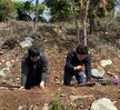 나무 앞에서 삽을 들고 식재 작업에 참여하고 있는 40대 남성과 여성. 배경에는 울창한 산과 나무들이 있으며, 두 사람은 밝게 웃으며 나무를 심고 있다. 여성은 아이보리색 재킷을, 남성은 하늘색 바람막이를 입고 있으며, 주변에는 함께 참여하는 사람들이 보인다. 오른쪽에는 세워진 바위 조형물도 있다.