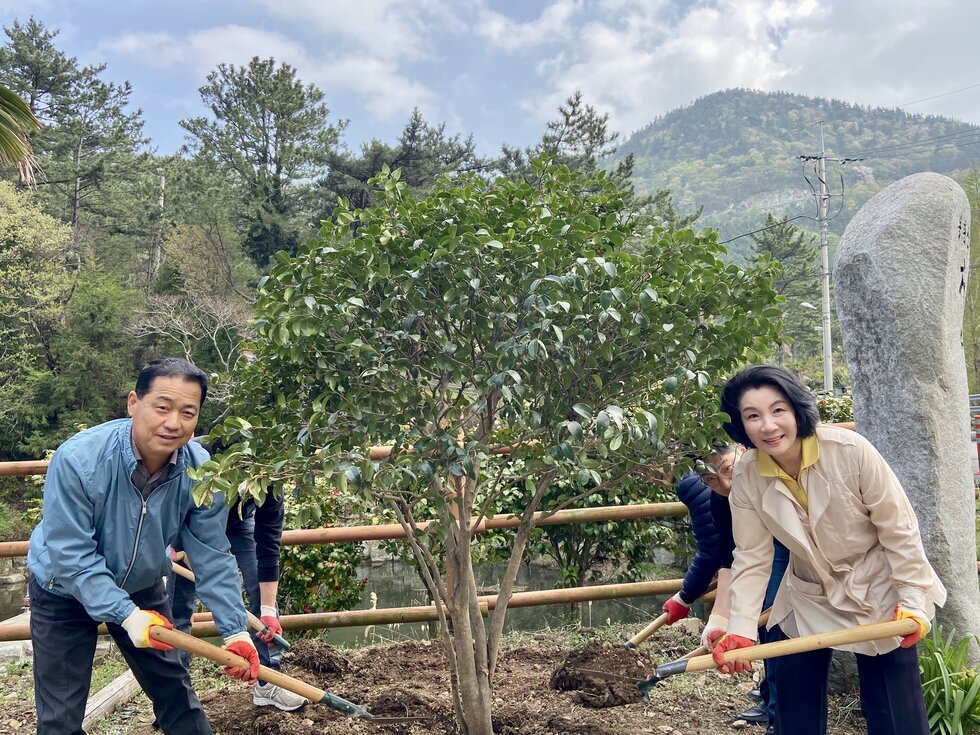 나무 앞에서 삽을 들고 식재 작업에 참여하고 있는 40대 남성과 여성. 배경에는 울창한 산과 나무들이 있으며, 두 사람은 밝게 웃으며 나무를 심고 있다. 여성은 아이보리색 재킷을, 남성은 하늘색 바람막이를 입고 있으며, 주변에는 함께 참여하는 사람들이 보인다. 오른쪽에는 세워진 바위 조형물도 있다.