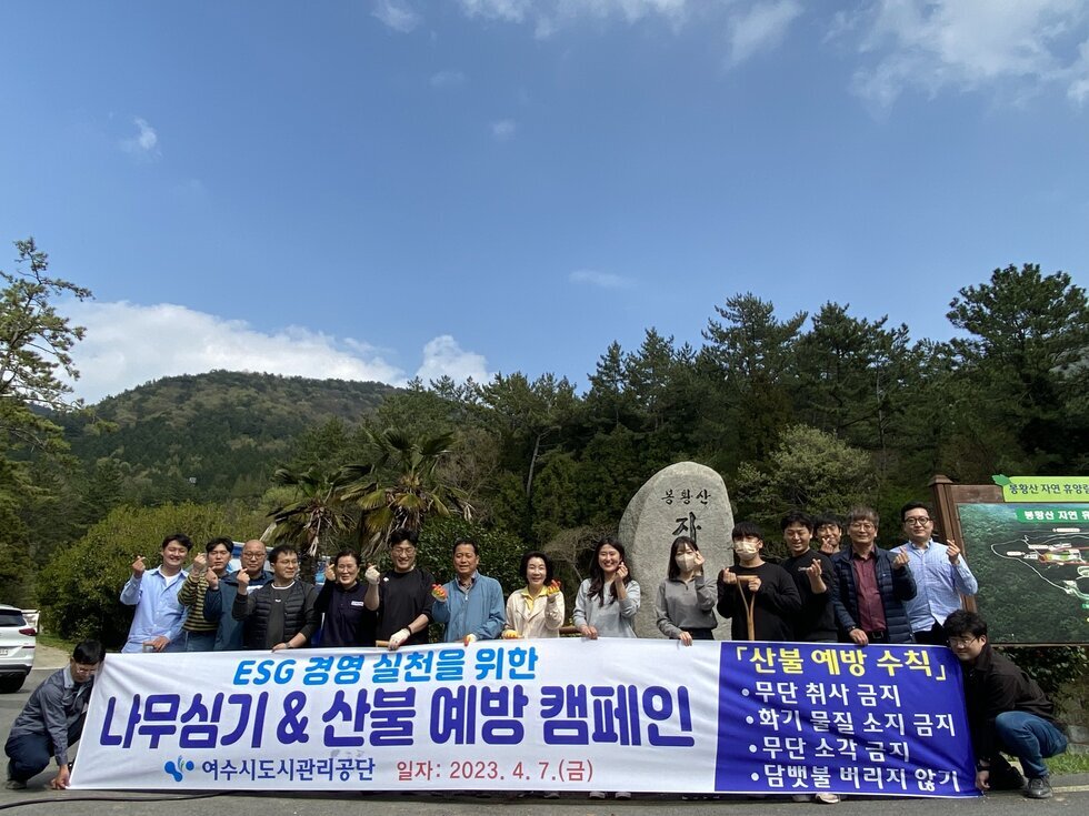 'ESG 경영 실천을 위한 나무심기 & 산불 예방 캠페인 여수시도시관리공단 일자 : 2023. 4. 7. (금) '이라는 문구가 적힌 현수막을 들고 20여 명의 여수시도시관리공단 직원들이 봉황산 입구에서 기념촬영을 하고 있다. 참가자들은 각자 장갑을 끼고 주먹을 쥐거나 손을 흔들며 캠페인 참여 의지를 나타내고 있다. 배경에는 산과 푸른 하늘, '봉황산'이라고 새겨진 바위가 함께 보이며 자연 친화적인 분위기를 더한다. 연령대는 20대부터 60대까지 다양하다.