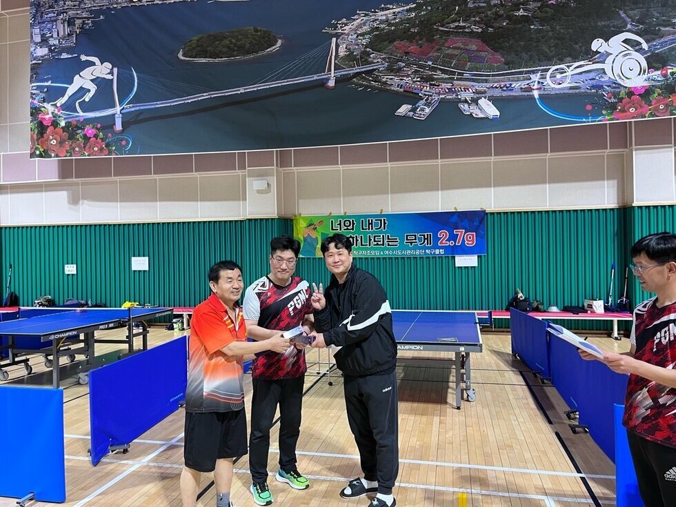 세 명의 남성이 탁구대 앞에서 밝은 표정으로 기념 촬영을 하고 있으며, 세명 모두 하나의 수상패를 함께 잡고 있다. 한 사람은 손가락으로 브이(V) 포즈를 취하며 활짝 웃고 있다. 오른편에는 종이를 든 남자가 이를 바라보며 환히 웃고있으며 배경에는 '너와 내가 하나되는 무게 2.7g'이라는 문구가 적힌 플랜카드가 걸려 있다.