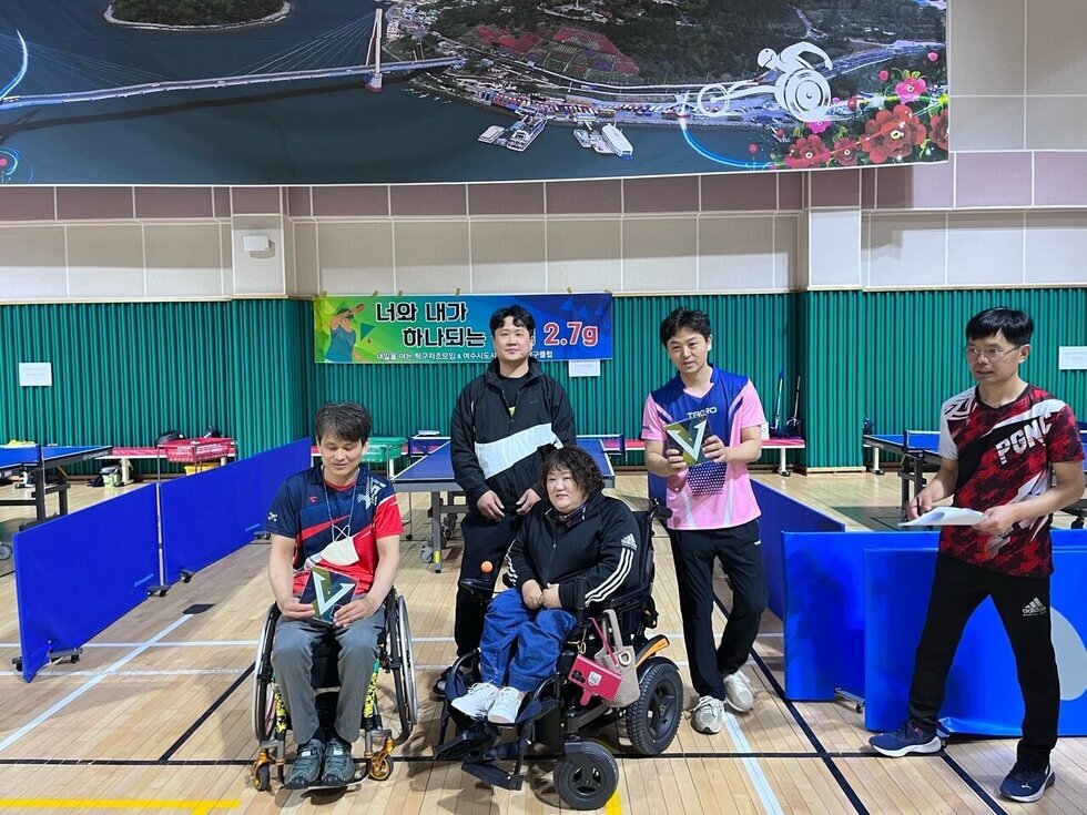 실내 체육관에서 휠체어를 탄 두 명의 남녀 참가자와 세 명의 일반 참가자들이 탁구대 앞에서 기념촬영을 하고 있다. 배경에는 ‘너와 내가 하나되는 무게 2.7g’이라는 플랜카드가 걸려있다. 참가자들은 탁구 대회 수상 기념패를 들고 환하게 웃으며 카메라를 응시하고 있다.