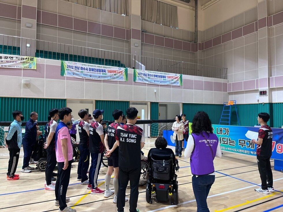 휠체어를 탄 참가자들과 자원봉사자, 관계자 등 20여 명이 배구 네트를 사이에 두고 한쪽을 바라보며 주의를 기울이고 있다. 오른쪽에는 진행자가 안내말을 전하고 있으며, 참가자들은 팀복을 입고 단체로 정렬되어 있다. 벽에는 '2022 전남 여수시 장애인생활체육 어울림배구교실 '이라고 적힌 현수막 등 다양한 체육 프로그램 홍보 배너가 걸려 있어 활기찬 분위기를 전달한다.