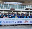'따뜻한 가치, 섬세한 서비스, 든든한 도시관리공단 여수시도시관리공단 노‧사 합동 동행 봉사단' 이라는 문구가 적힌 대형 현수막을 펼쳐 들고, 그 뒤로는 '아름다운 나눔보따리 희망을 배달합니다.' 등의 문구가 적힌 현수막이 붙은 여수시청 앞 계단에서 기념촬영한 모습이다. 여수 시청 건물에는 여수시 남해안 거점도시 미항 여수라는 현수막이 걸려있다. 생필품이 담긴 나눔 보따리를 전달하기 위해 모인 여수시도시관리공단 봉사단 50여 명이 단체로 서 있으며, 대체로 20대에서 50대 연령대의 직원들로 구성되어 있다. 일부는 손가락 하트나 엄지손가락을 들어 보이며 포즈를 취하고 있다.