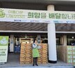'따뜻한 가치, 섬세한 서비스, 든든한 도시관리공단 여수시도시관리공단 노‧사 합동 동행 봉사단' 이라는 문구가 적힌 대형 현수막을 펼쳐 들고, 그 뒤로는 '아름다운 나눔보따리 희망을 배달합니다.' 등의 문구가 적힌 현수막이 붙은 여수시청 앞 계단에서 기념촬영한 모습이다. 여수 시청 건물에는 여수시 남해안 거점도시 미항 여수라는 현수막이 걸려있다. 생필품이 담긴 나눔 보따리를 전달하기 위해 모인 여수시도시관리공단 봉사단 50여 명이 단체로 서 있으며, 대체로 20대에서 50대 연령대의 직원들로 구성되어 있다. 일부는 손가락 하트나 엄지손가락을 들어 보이며 포즈를 취하고 있다.