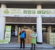 '따뜻한 가치, 섬세한 서비스, 든든한 도시관리공단 여수시도시관리공단 노‧사 합동 동행 봉사단' 이라는 문구가 적힌 대형 현수막을 펼쳐 들고, 그 뒤로는 '아름다운 나눔보따리 희망을 배달합니다.' 등의 문구가 적힌 현수막이 붙은 여수시청 앞 계단에서 기념촬영한 모습이다. 여수 시청 건물에는 여수시 남해안 거점도시 미항 여수라는 현수막이 걸려있다. 생필품이 담긴 나눔 보따리를 전달하기 위해 모인 여수시도시관리공단 봉사단 50여 명이 단체로 서 있으며, 대체로 20대에서 50대 연령대의 직원들로 구성되어 있다. 일부는 손가락 하트나 엄지손가락을 들어 보이며 포즈를 취하고 있다.