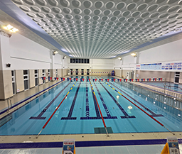 4개의 레인이 구분된 실내 수영장 전경으로, 천장에는 원형 패턴의 구조물이 설치되어 있고 우측 벽면에는 이용 안내 및 안전 수칙 표지판이 부착된 쾌적한 수영 시설 모습