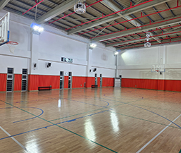 넓은 우드 플로어링 바닥과 높은 층고를 갖춘 실내 체육관으로, 벽면 하단에는 붉은색 안전 보호대가 설치되어 있으며 농구 골대와 전자 점수판이 구비된 다목적 체육 시설 전경