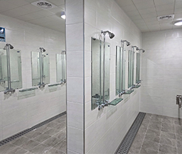 화이트 톤의 타일 벽면과 그레이 톤의 미끄럼 방지 바닥으로 마감된 샤워실 내부에 샤워기, 거울, 비누 거치대가 일정한 간격으로 설치된 청결한 샤워 시설 모습