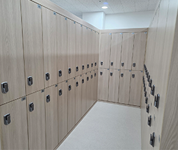 밝은 우드 톤의 개인 사물함이 벽면을 따라 정렬되어 있고, 각 사물함마다 잠금장치 핸들이 부착되어 있는 쾌적하고 깔끔한 탈의실 내부 전경