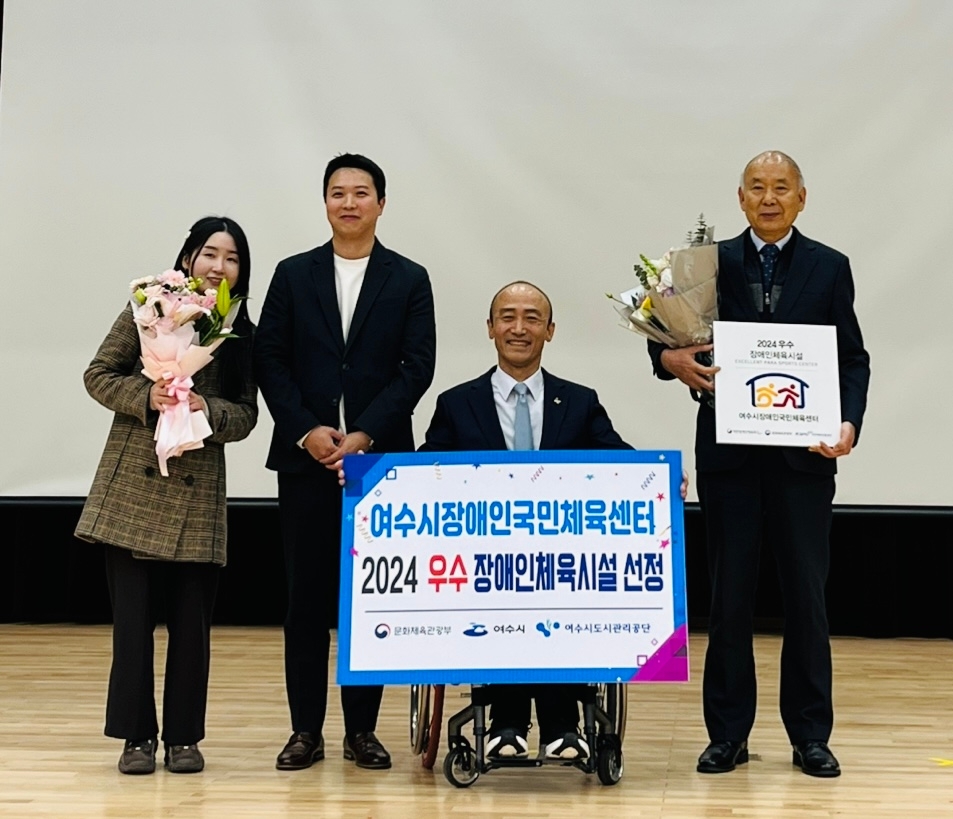 여수시장애인국민체육센터 2024 우수 장애인체육시설 선정 문화체육관광부 여수시 여수시도시관리공단 팜플렛을 들고 있는 남성이 휠체어에 타고 있고 양 옆으로 분홍색 꽃다발을 들고 있는 여성, 검은색 자켓을 입고 있는 남성과, 꽃다발과 2024 우수 장애인체육시설 인증 팻말을 들고 있는 남성이 보인다