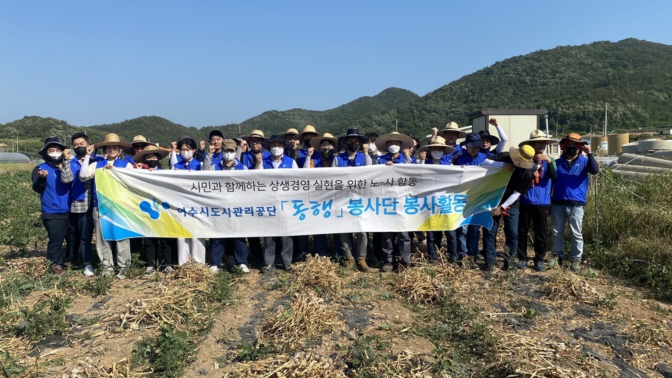 시민과 함께하는 상생경영 실현을 위한 노-사 합동 여수시도시관리공단 동행 봉사단 봉사활동 이라고 적힌 현수막을 들고 봉사단원들이 마늘밭에서 기념촬영을 하는 모습