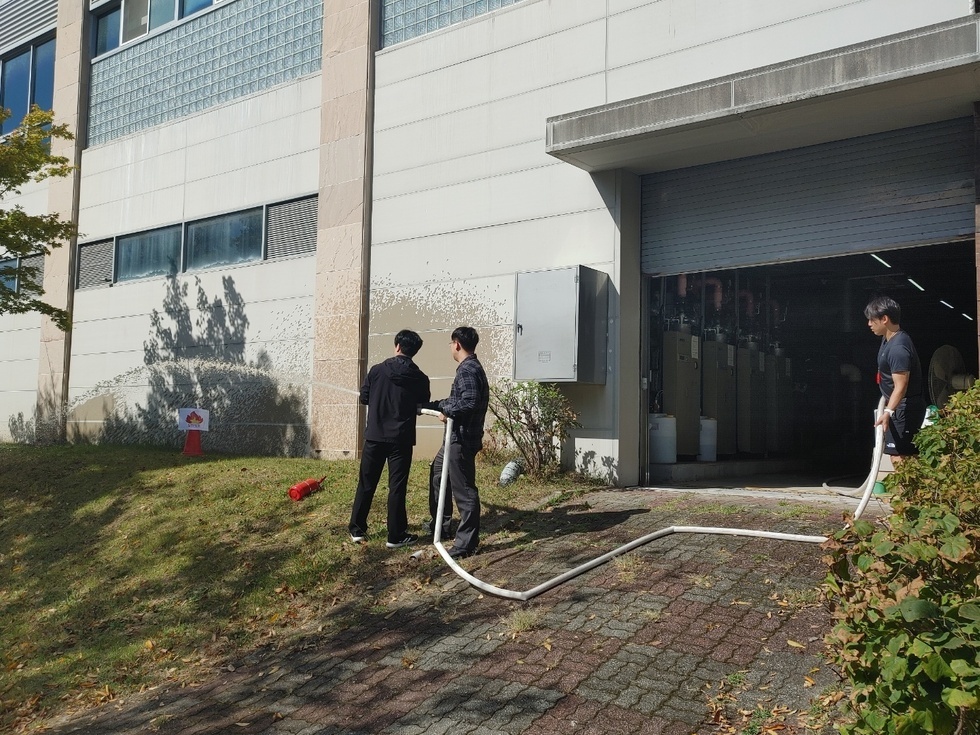 건물 앞에서 3명의 사람들이 소방 호스를 들고 화단에 물을 뿌리고 있는 모습