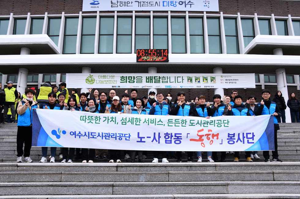 따뜻한 가치, 섬세한 서비스, 든든한 도시고나리공단 / 여수시도시관리공단 노-사 합동 동행 봉사단 이라고 적힌 큰 현수막을 파란색 옷을 입은 사람들이 들고 계단위에서 사진을 찍고 있는 모습