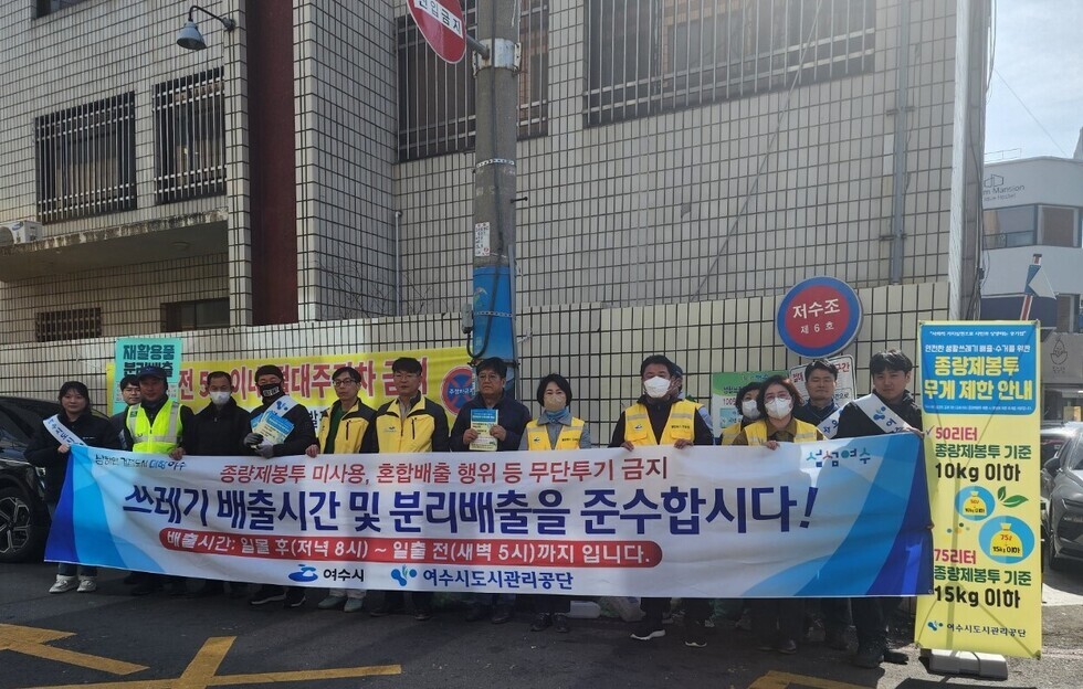 종량제봉투 미사용, 혼합배출 행위 등 무단 투기 금지 / 쓰레기 배출 시간 및 분리 배출을 준수 합시다/ 배출 시간 : 일몰 후(저녁 8시) ~ 일출 전 (새벽 5시)까지 입니다 / 여수시, 여수도시관리공단 이라고 적힌  큰 현수막을 노란색 옷을 입은 사람들이 들고 있는 사진