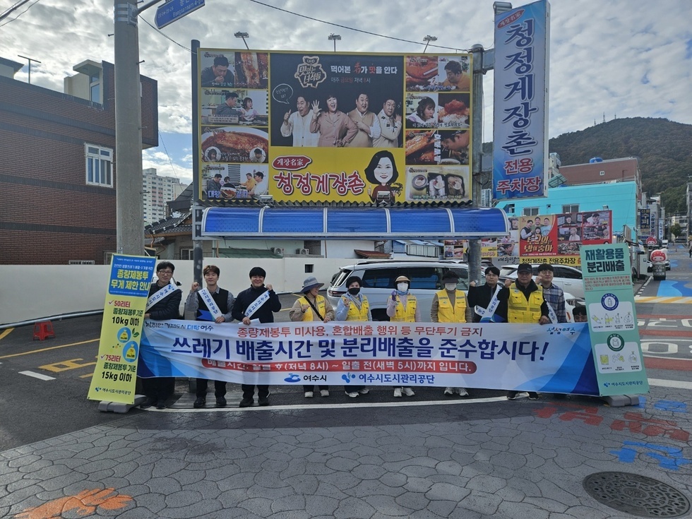 청정게장촌 주차장 앞에서 쓰레기 분리배출을 홍보하는 캠페인에 참여한 여수시와 여수시도시관리공단 관계자들이 현수막(종량제봉투 미사용, 혼합배출 행위 등 무단투기금지 쓰레기 배출시간 및 분리배출을 준수합시다! 배출시간 일몰 후 (저녁 8시)~일출전(새벽5시)까지 입니다. 여수시,여수시도시관리공단)을 들고 화이팅 포즈로 기념촬영을 하는 모습