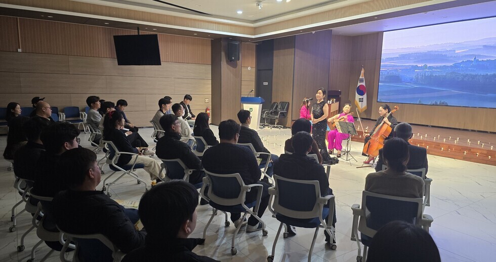 여러명의 직원들이 의자에 앉아있고 직원들 앞에서 검은색 드레스를 입은 중년여성이 노래를 부르고 그 뒤에는 연주자들이 각각 악기를 연주하는 모습을 찍은 사진이다.