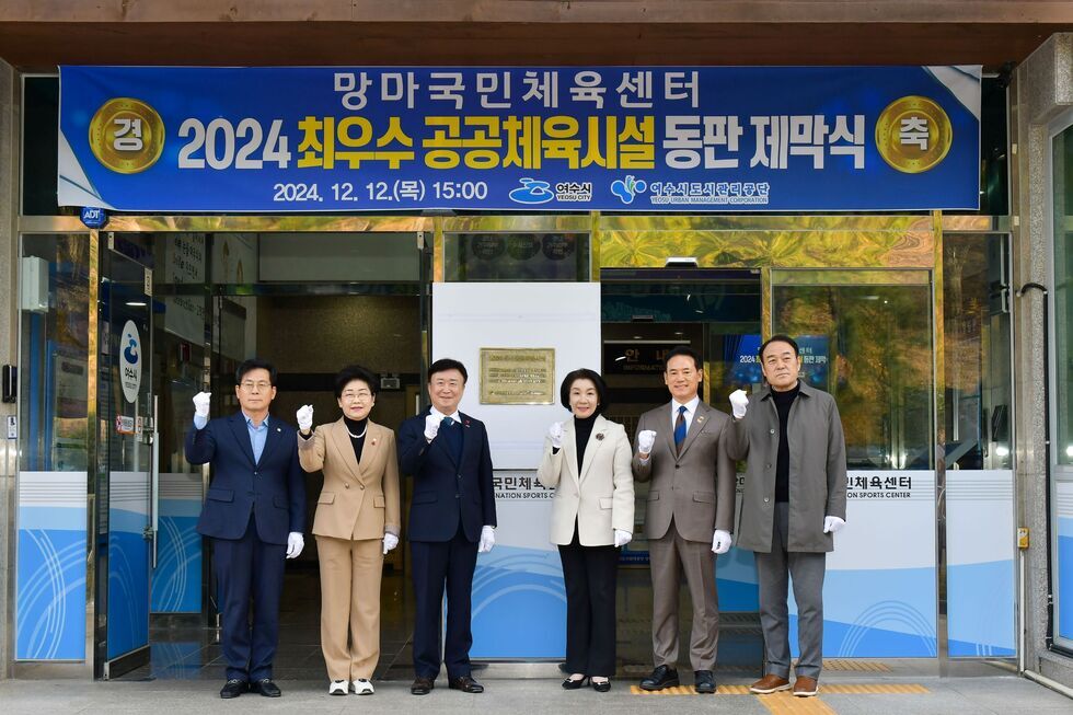 '망마국민체육센터경 2024 최우수 공동체육시설 동판 제막식 축2024. 12. 12.(목) 15:00 여수시 여수시도시관리공단' 문구가 적힌 현수막이 걸려있는 건물 앞에서 여수도시관리공단 김유화 이사장 여수시장, 관계자들의 기념촬영 모습