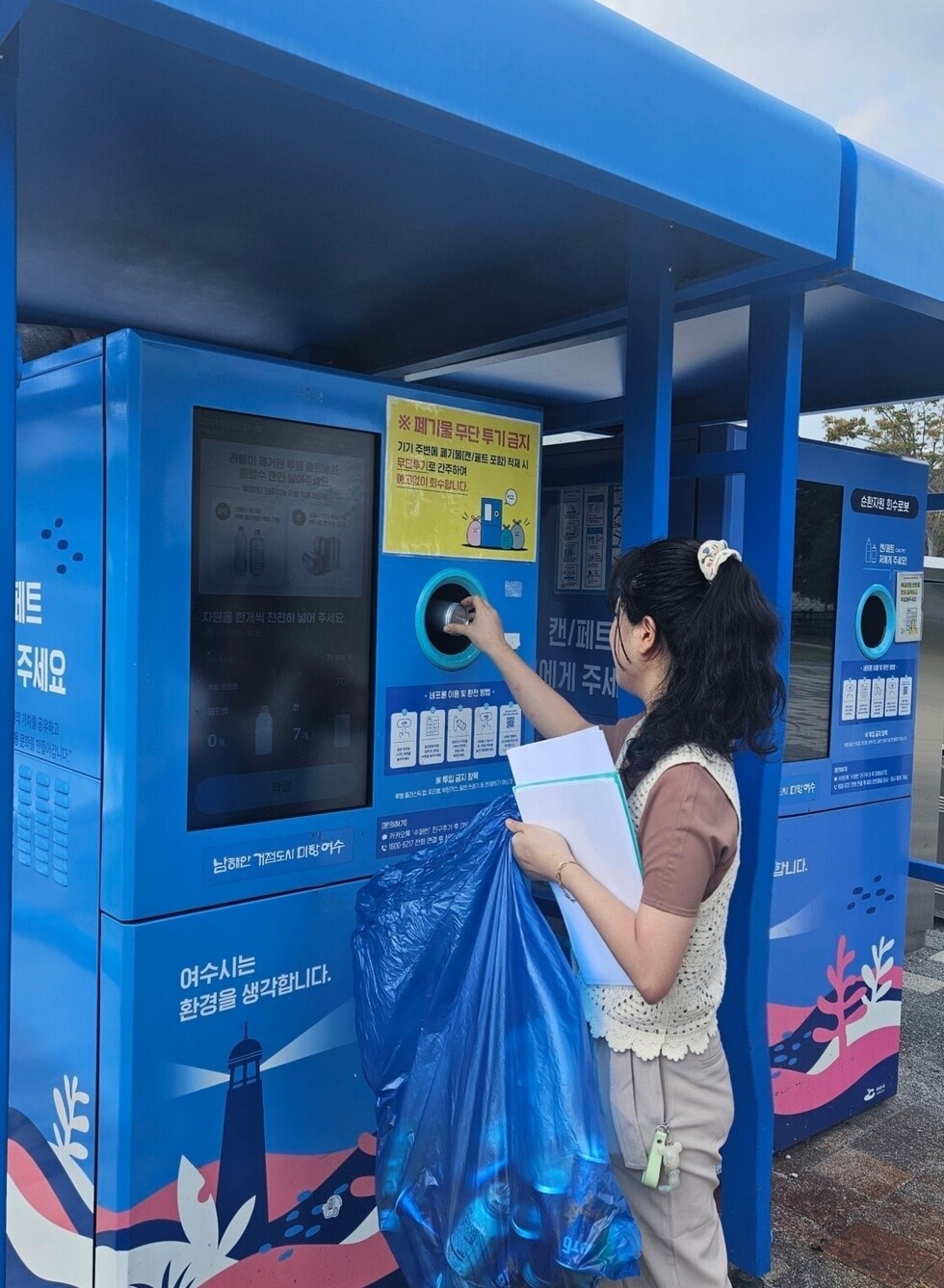 여성시민이 재활용 캔이 많이 든 봉지를 들고 페트병을 자동으로 수거하는 인공지능 자동수거기에 캔을 넣고 있는 모습