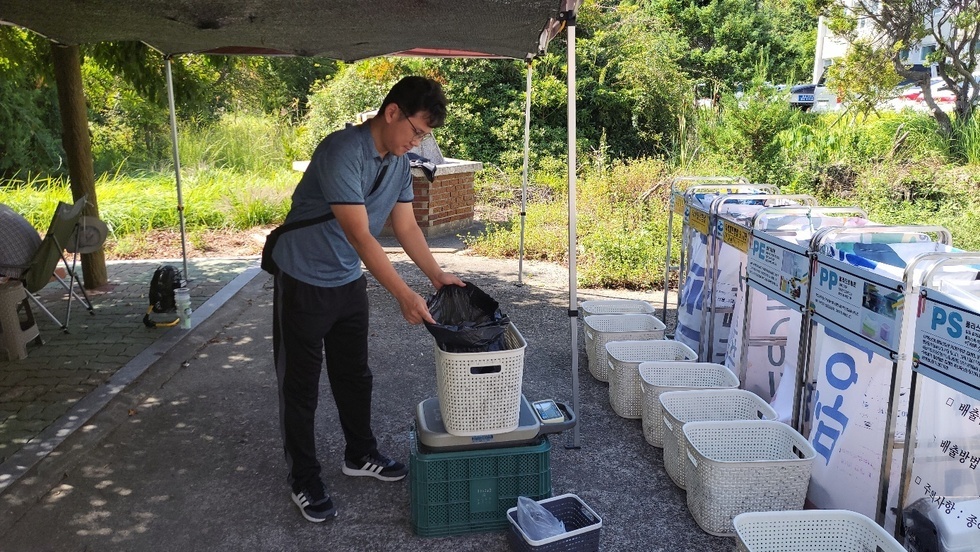 한 중년남성이 흰 바구니 안에 검은색 봉투를 넣어서 저울 위에 올려 무게를 재고 있는 모습이다. 앞에는 여러 개의 분리수거함이 존재하며 앞에는 여러개의 바구니가 배치되어있는 사진이다. 한 분리수거함 안의 봉투에는 '배출, 배출방법, 주의사항' 문구가 적혀있다.