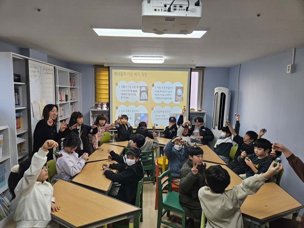 지역아동센터 학생 대상으로 폐셔틀콕 키링 제작 과정에 대해 교육하고 있다. 키링 제작 과정의 4단계가 뒷 배경화면에 뛰워져 있고 키링을 들고 자랑하고 있는 아이들의 사진