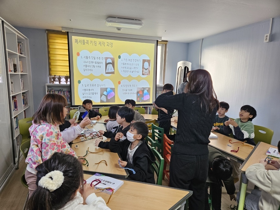 지역아동센터 학생 대상으로 폐셔틀콕 키링 제작 과정에 대해 교육하고 있다. 상하의 검은색 옷을 입은 여성이 아이들 중심에 서서 폐셔틀콕 키링 제작 과정에 대해서 설명하고 있다