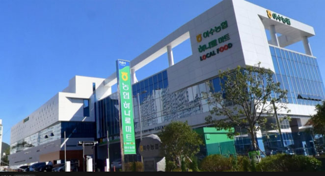 맑은 하늘 아래 위치한 현대적인 건물 외관. 건물 전면에는 '여수농협 하나로마트 LOCAL FOOD'라는 간판이 걸려 있으며, 중앙에는 유리창으로 된 입구가 있다. 입구 옆에는 초록색 배경에 흰 글씨로 된 농협·· 하나로마트 안내 간판이 세워져 있고, 주변에는 나무와 식물이 심어진 작은 녹지가 있다.