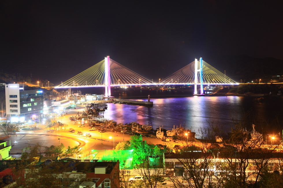 아름다운 조명이 더해진 여수의 거북선대교 야경. 형형색색의 불빛이 다리의 구조물을 따라 빛나며, 다리 아래 바다에는 조명이 반사되어 환상적인 분위기를 연출한다. 전경에는 불빛이 가득한 항구와 도심의 거리, 정박된 선박들이 보이며, 배경에는 어둠 속 산이 실루엣처럼 자리하고 있다.