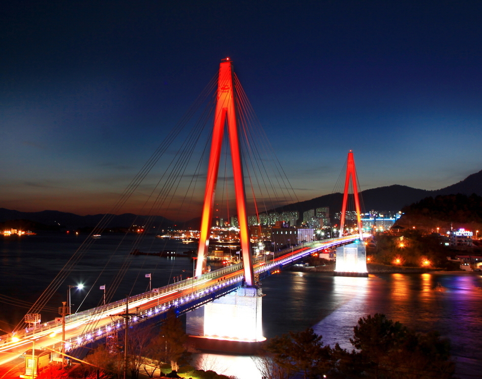 붉은 조명이 아름답게 비춰진 돌산대교의 야경. 다리는 푸른 바다 위를 가로지르며 도시의 불빛과 어우러져 화려한 분위기를 자아낸다. 배경에는 어두운 하늘과 산자락이 어렴풋이 보이며, 다리 아래 수면에는 조명빛이 반사되어 반짝인다.