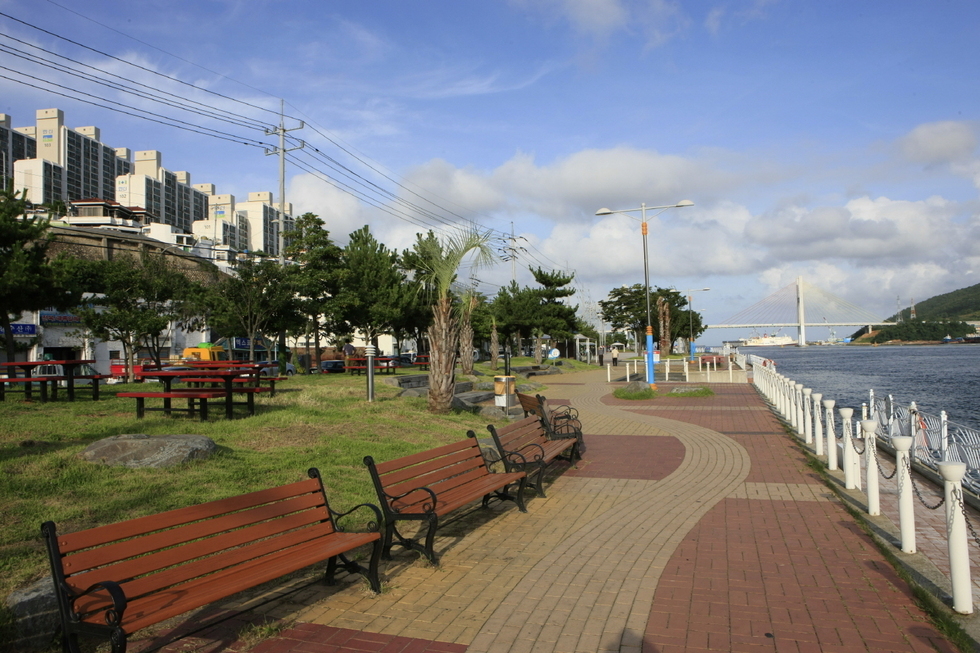 종포해양공원 산책로의 모습. 오른쪽으로는 푸른 바다가 펼쳐져 있고, 해안선을 따라 하얀 난간과 붉은 벽돌길이 이어진다. 왼쪽에는 나무 벤치와 잔디밭이 있고, 그 너머로 아파트 단지가 보인다. 멀리에는 다리가 보이며, 전체적으로 맑은 하늘과 구름이 어우러져 여유롭고 평화로운 분위기를 자아낸다.