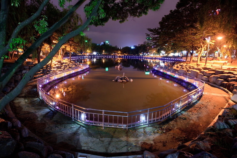 거북선 공원의 야경. 중앙에 조용한 연못이 자리하고 있으며, 연못 주변은 울타리와 보라색 조명으로 아름답게 장식되어 있다. 양옆으로는 키 큰 나무들이 우거져 있고, 오른쪽 산책로에는 가로등 불빛이 따뜻하게 공원을 비추고 있다. 조용하고 낭만적인 분위기 속에서 도시의 불빛이 멀리 반짝인다.