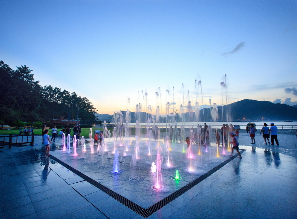 오동도의 여름 저녁, 분수광장에서 알록달록한 조명이 켜진 분수들이 시원하게 솟구치고 있다. 분수 주위에는 아이들과 가족들이 모여 시원한 물놀이를 즐기고 있고, 일부 사람들은 주변 벤치와 바다를 배경으로 풍경을 감상하고 있다. 배경에는 석양이 지는 하늘과 산, 바다가 어우러져 고요하고 아름다운 분위기를 자아낸다.