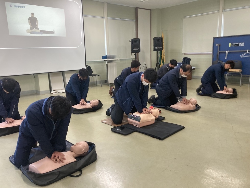8명의 사람들이 강의실 바닥에 무릎을 꿇고 CPR 마네킹을 대상으로 심폐소생술 교육을 받고 있다. 각자 앞에 놓인 마네킹에 압박을 가하며 실습 중이며, 뒤편 스크린에는 강사가 CPR 시범을 보이는 영상이 재생되고 있다. 참가자들은 동일한 네이비 작업복을 착용하고 있으며 마스크를 착용하고 있다.