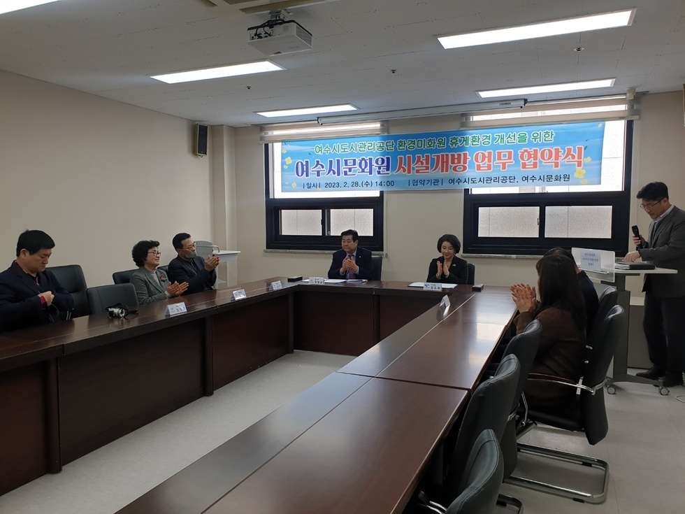 회의실에서 8명의 관계자들이 협약식 시작을 알리며 박수를 치고 있다. 정면 벽에는 ‘여수시도시관리공단 환경미화원 휴게환경 개선을 위한 여수시문화원 시설개방 업무 협약식 |일시| 2023. 2. 28 (수) 14:00 |협약기관| 여수시도시관리공단, 여수시문화원’ 이라고 적힌 협약식 현수막이 걸려 있으며, 책상에는 이름표가 놓여 있다.  가운데 위치한 50대 남, 녀를 중심으로 양쪽으로 앉은 참석자들이 보이며 오른쪽에는 한명의 진행자가 마이크를 들고 있는 모습이다. 