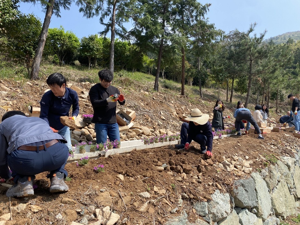 꽃모종을 심기 위해 흙을 고르고 돌을 치우는 10여명의 사람들이 있으며, 앞쪽에 모자를 쓴 사람은 쪼그려 앉아 땅을 고르고 있다. 주변에는 상자 속 꽃모종들이 놓여 있고, 뒷편에는 나무들이 울창하게 자라난 산림이 펼쳐져 있다.