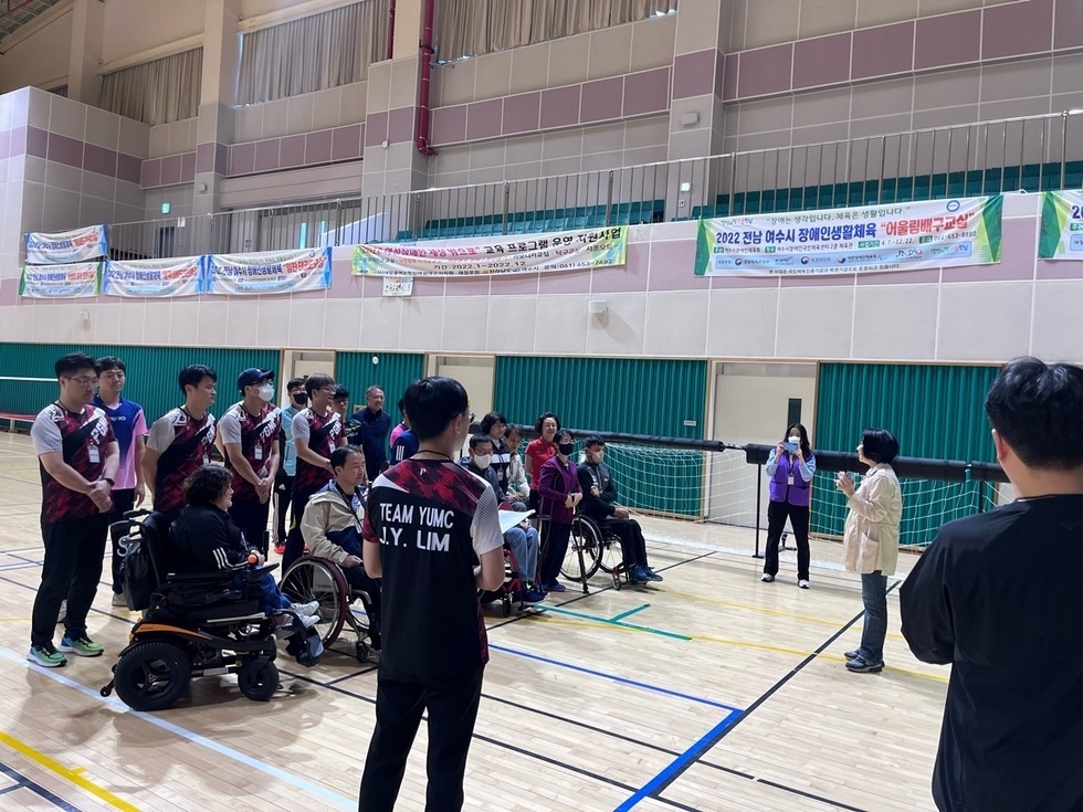 휠체어를 탄 참가자들과 자원봉사자, 관계자 등 20여 명이 배구 네트를 사이에 두고 한쪽을 바라보며 주의를 기울이고 있다. 오른쪽에는 진행자가 안내말을 전하고 있으며, 참가자들은 팀복을 입고 단체로 정렬되어 있다. 벽에는 '2022 전남 여수시 장애인생활체육 어울림배구교실 '이라고 적힌 현수막 등 다양한 체육 프로그램 홍보 배너가 걸려 있어 활기찬 분위기를 전달한다.