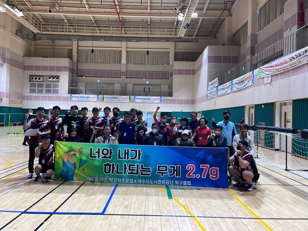 실내 체육관에서 '너와 내가 하나되는 무게 2.7g 내일을 여는 탁구자조모임 & 여수시도시관리공단 탁구클럽'이라는 문구가 적힌 현수막을 들고 단체 기념사진을 촬영하는 참가자들. 약 30여 명이 참여하였으며, 휠체어 이용자와 비장애인이 함께 어우러져 손을 들고 밝은 표정으로 포즈를 취하고 있다. 배경에는 현수막들이 걸려 있고, 실내 조명 아래 훈훈한 분위기가 느껴진다.